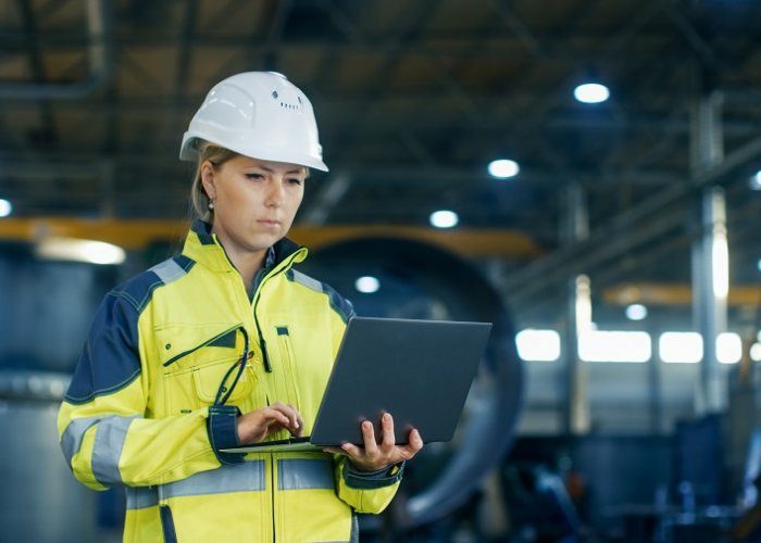 Female Industrial Engineer in the Hard Hat Uses Laptop Computer while Standing in the Heavy Industry Manufacturing Factory. In the Background Various Metalwork Project Parts Lying